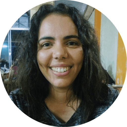 Foto da Alana Bacco. Mulher, branca, cabelos escuros, cacheados e compridos, vestindo uma camiseta cinza e sorrindo.